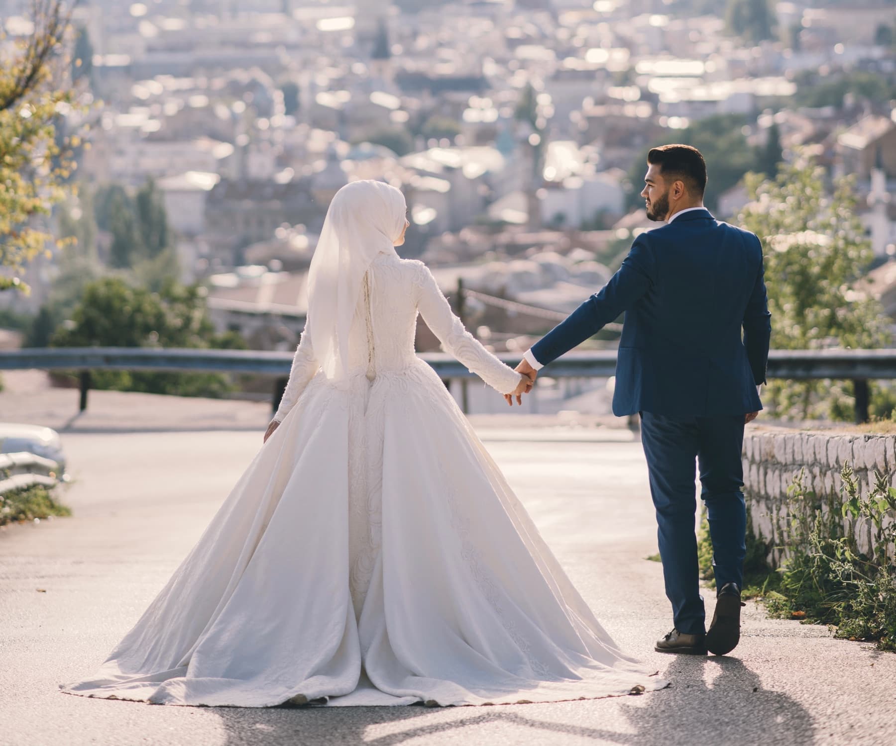 A bride and groom walking hand in hand along a road above a hillside city